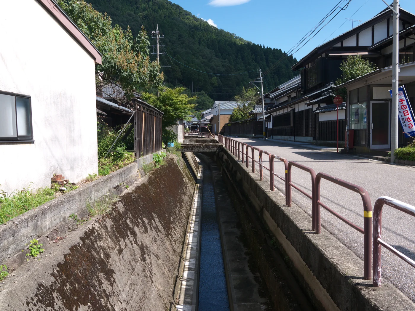 越前和紙之里街區的水道、欄杆與黑白外牆家屋,後方是樹林覆蓋的山坡