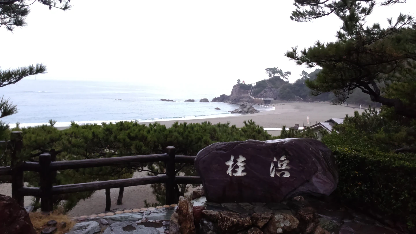 桂濱石碑位在松樹與欄杆前，後方可見弧形海灣與龍王岬