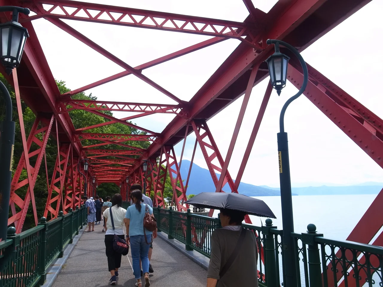 遊客步行通過支笏湖山線鐵橋,紅色桁架旁可見湖面