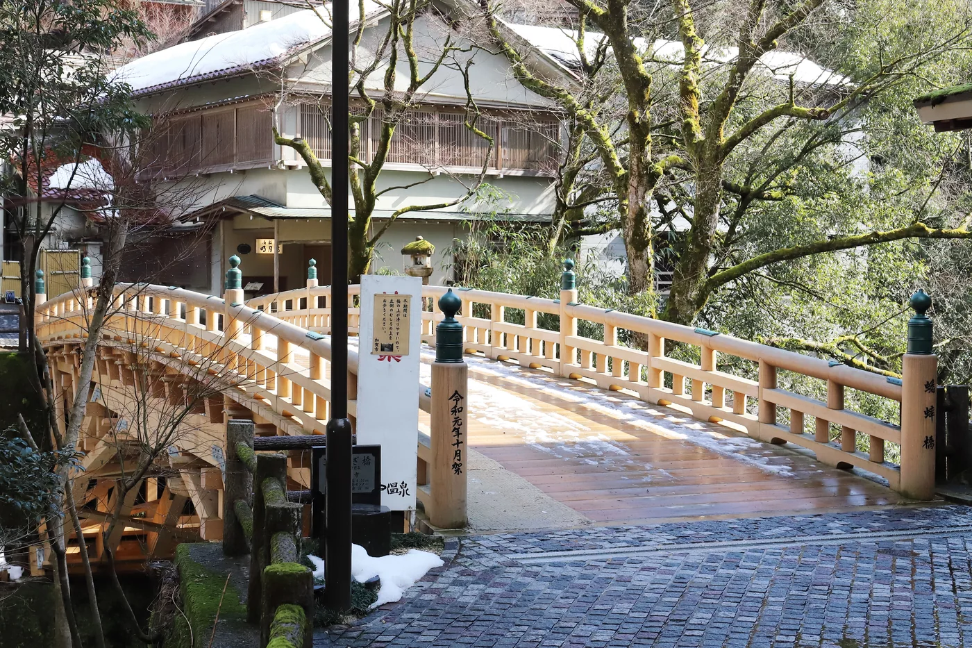 山中溫泉こおろぎ橋的淺色木欄杆與殘雪,前方是濕潤石板路,周圍有樹木