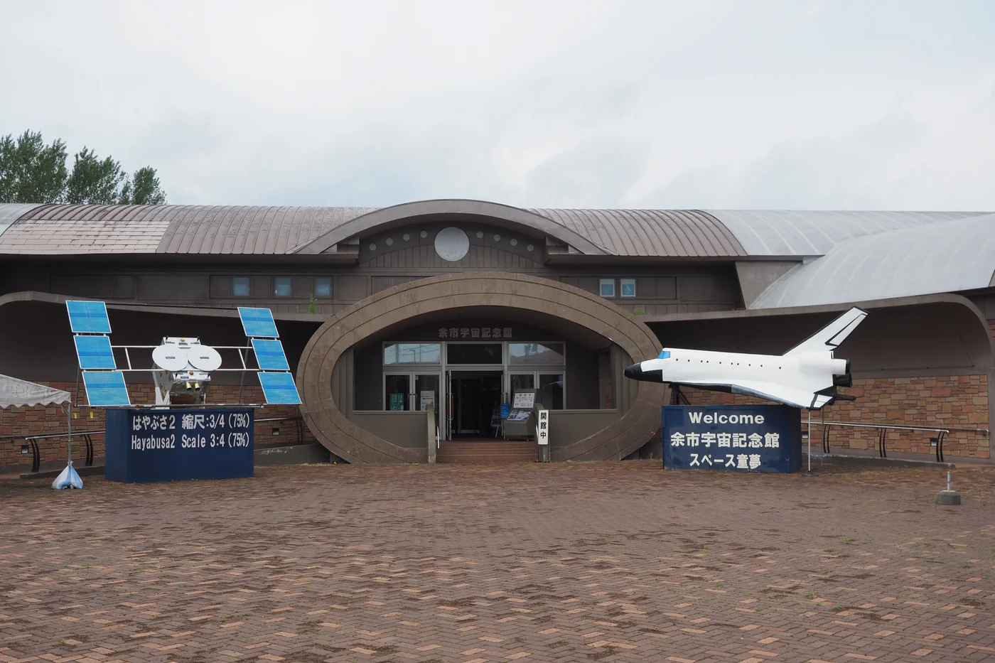 余市宇宙記念館褐色館舍正面，中央是橢圓形入口，左側擺放帶藍色太陽能板的探測器模型，右側有白色太空梭模型